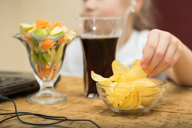 Une fille mange des croustilles et une salade de fruits devant un ordinateur portable.