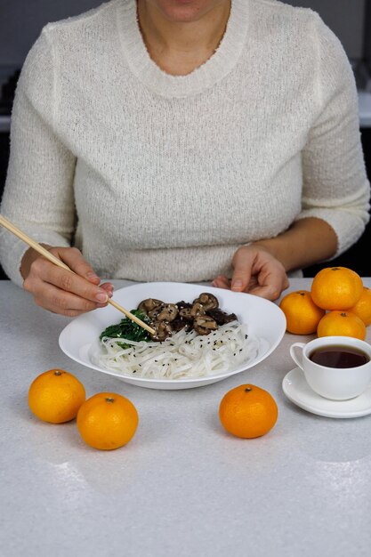 La fille mange dans une assiette avec des nouilles de riz et des champignons shiitake Mug blanc avec du café et des mandarines au premier plan
