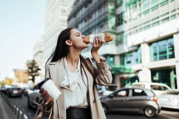 La fille mange avec impatience un hamburger en déplacement dans le contexte des grands immeubles