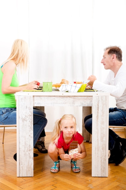 Fille mangeant du chocolat sous la table pendant que la famille prend son petit déjeuner