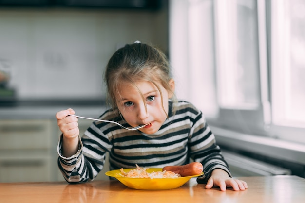 Fille de manger des pâtes avec des saucisses dans la cuisine dans une veste rayée