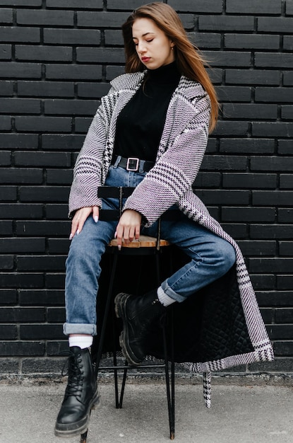 une fille en manteau est assise sur une chaise près d'un mur noir