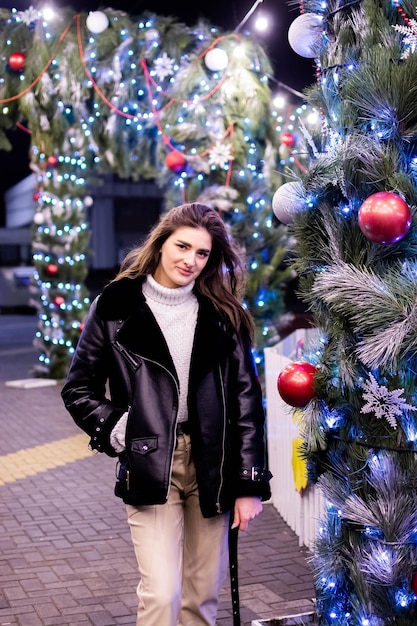Une fille en manteau et gants se tient sous un sapin de Noël décoré de lumières. Nouvel An 2022.
