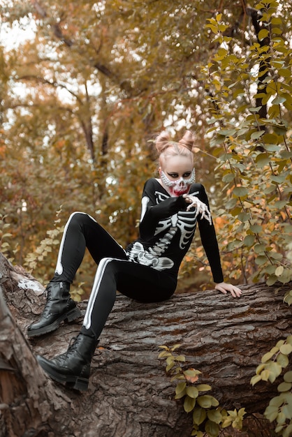 Une fille avec un maquillage effrayant dans un costume de squelette est assise sur un vieil arbre desséché. Halloween