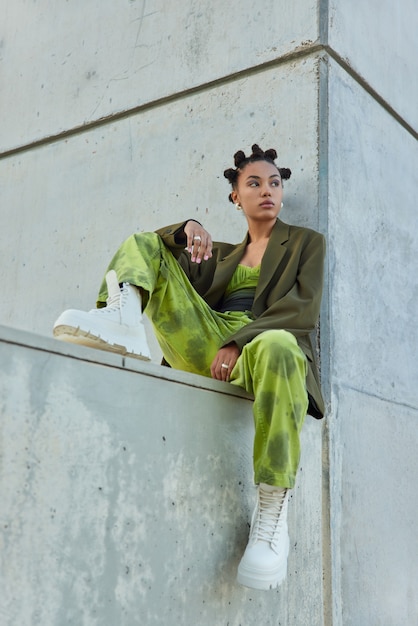 fille avec un maquillage lumineux vêtue de vêtements verts à la mode des bottes blanches pose contre un mur gris regarde loin passe son temps libre en milieu urbain