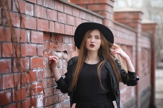 Une fille marche dans la rue dans un gilet en cuir avec un téléphone. Jeune belle fille dans un chapeau et avec un maquillage sombre à l'extérieur. Fille de style gothique dans la rue.