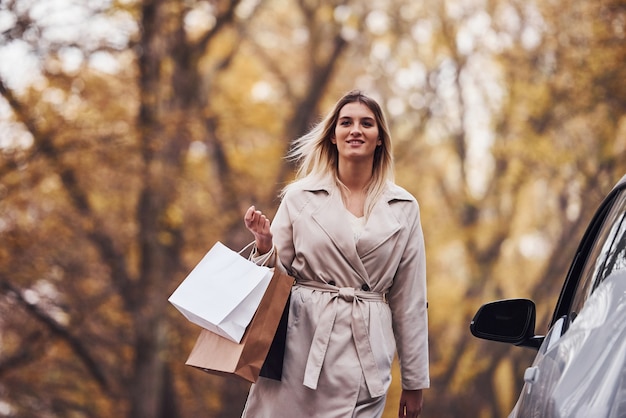 Fille marche près de la voiture avec des sacs en mains. Nouvelle automobile moderne dans la forêt.