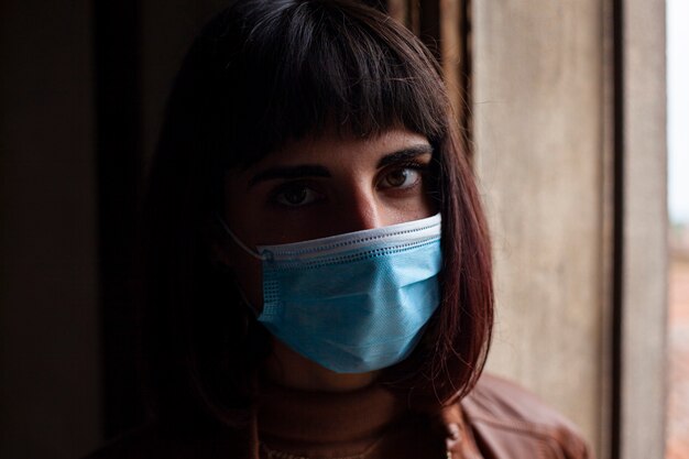 Fille avec masque médical à la fenêtre de sa maison pendant la période de quarantaine covid