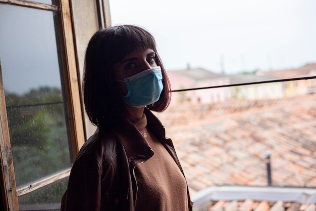 Photo fille avec masque médical à la fenêtre de sa maison pendant la période de quarantaine covid