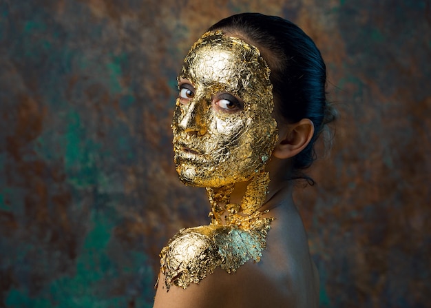 Fille avec un masque sur son visage fait de feuille d'or Portrait en studio sombre d'une brune sur un résumé