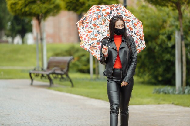 fille masquée marche dans la rue. fille dans un masque de protection se promène dans le parc avec un parapluie sous la pluie. Infection à coronavirus COVID-19