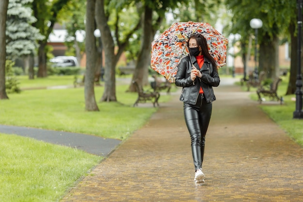 fille masquée marche dans la rue. fille dans un masque de protection se promène dans le parc avec un parapluie sous la pluie. Infection à coronavirus COVID-19