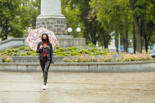 Une fille masquée marche le long de la rue. Une fille dans un masque de protection se promène dans le parc avec un parapluie sous la pluie. Infection à coronavirus COVID-19.