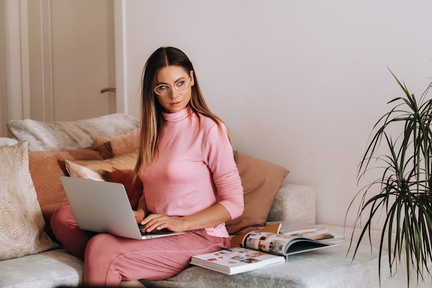 Une fille le matin en pyjama et lunettes à la maison travaille sur un ordinateur portable, une fille s'auto-isole à la maison et se repose sur le canapé et regarde un ordinateur portable. Tâches ménagères