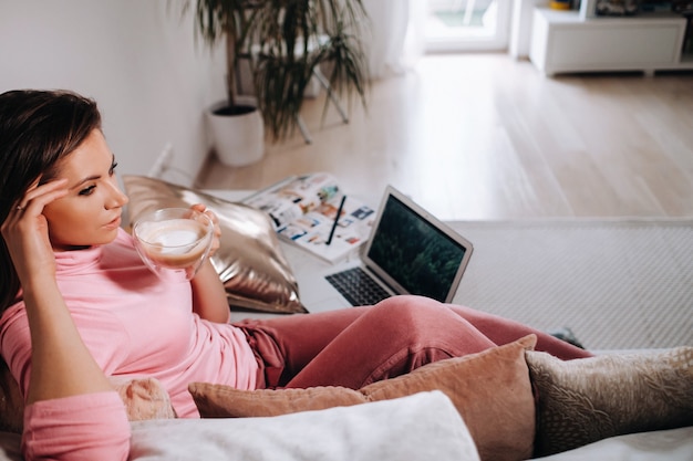 Une fille le matin en pyjama à la maison travaillant sur un ordinateur portable en buvant du café, une fille s'isolant à la maison et se reposant sur le canapé et regardant un ordinateur portable. Tâches ménagères