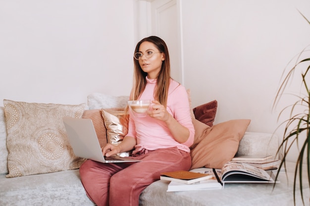 Une fille le matin en pyjama à la maison travaillant sur un ordinateur portable avec du café, une fille isolée à la maison et se reposant sur le canapé et regardant un ordinateur portable.