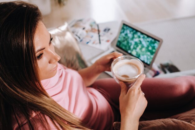 Une fille le matin en pyjama à la maison travaillant sur un ordinateur portable avec du café, une fille isolée à la maison et se reposant sur le canapé et regardant un ordinateur portable.