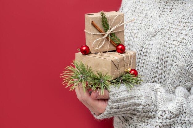 Une fille méconnaissable en pull tient des cadeaux de Noël sur fond rouge. Coffrets cadeaux faits à la main dans les mains d'une femme. Fond de nouvel an avec de beaux cadeaux.