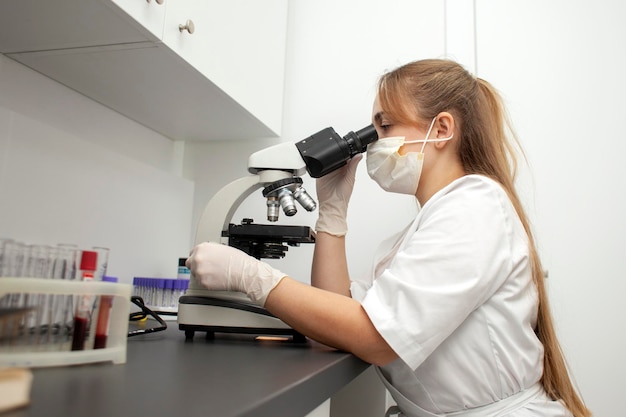 une fille médecin en uniforme et masque médical examine le microscope dans le laboratoire