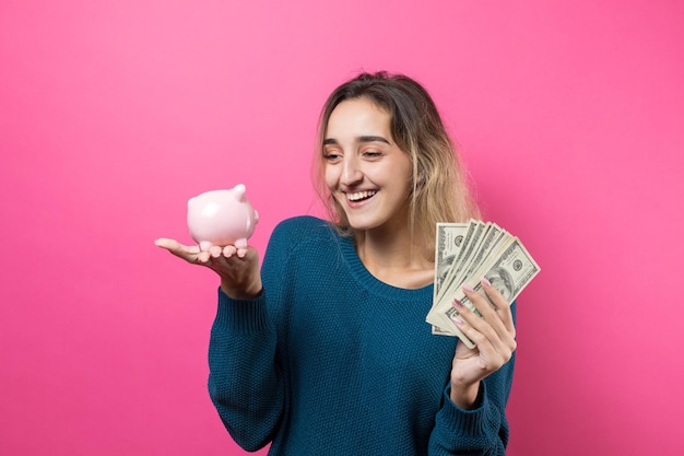 Fille met dans la tirelire des billets de cent dollars Jeune fille sur fond rose tenant une tirelire et se réjouit en faisant des gestes Le concept de fiabilité des investissements en espèces et de l'assurance