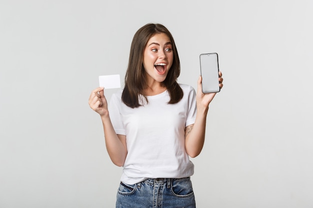 Fille mignonne excitée et surprise montrant la carte de crédit et l'application bancaire de téléphone mobile à l'écran.