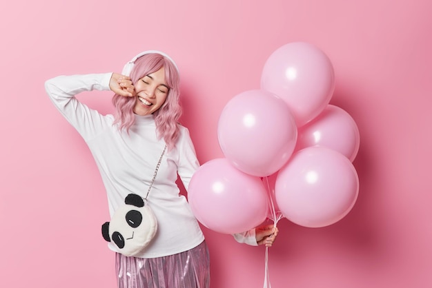 Une fille millénaire aux cheveux longs positive écoute sa musique préférée tient un tas de ballons gonflés porte un sac porte un col roulé blanc et une jupe a une ambiance festive isolée sur fond rose Concept de vacances