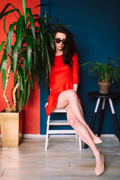 Fille mince belle mode aux cheveux longs noirs, en robe élégante rouge et lunettes de soleil posant sur le mur rouge bleu.