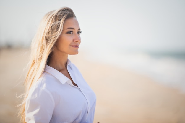 Une fille mince bronzée dans un maillot de bain bleuâtre et une chemise blanche légère, profite d'un été chaud et ensoleillé sur la côte d'une mer bleue déchaînée