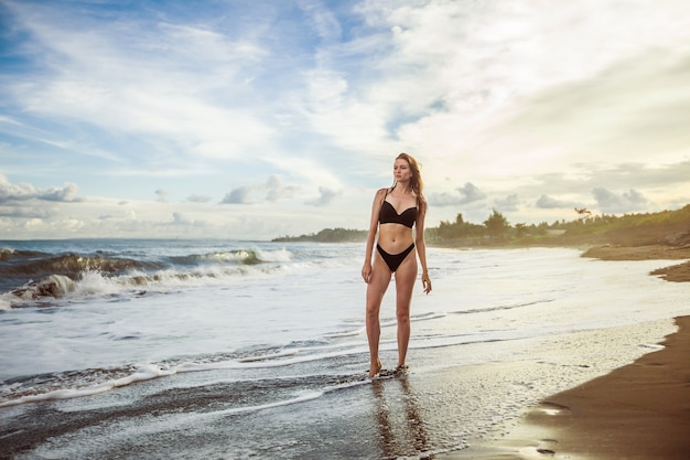 Fille mince dans un maillot de bain noir est debout sur une plage dans le contexte d'un beau coucher de soleil