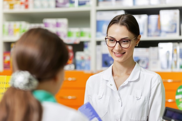 Une fille mince avec de longs cheveux noirs portant un manteau blanc parle avec un visiteur dans une pharmacie