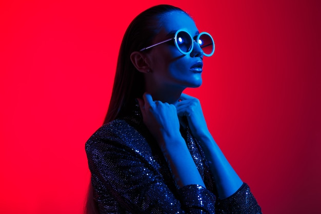 Fille de mode aux cheveux longs et lunettes de soleil rondes dans une robe noire brillante pose à la lumière du néon dans le studio.