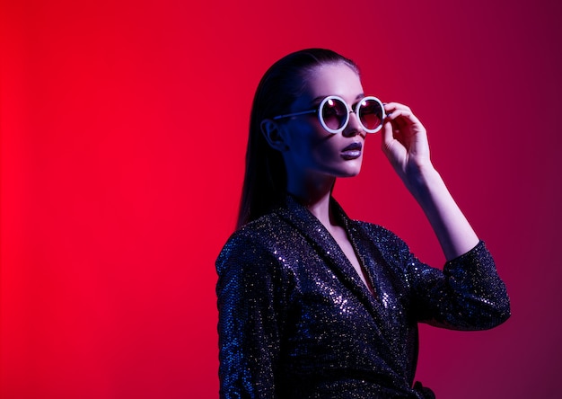 Fille de mode aux cheveux longs et lunettes de soleil rondes dans une robe noire brillante pose à la lumière du néon dans le studio.