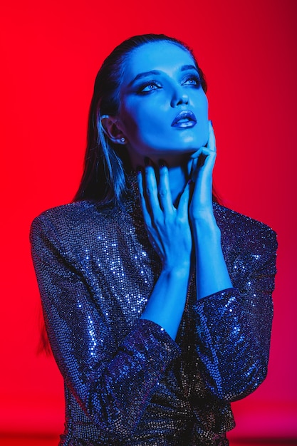 Fille de mode aux cheveux longs et maquillage élégant dans une robe noire brillante pose sur fond rouge à la lumière du néon dans le studio