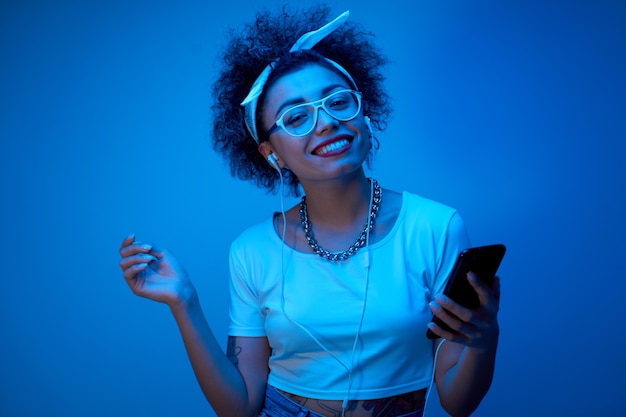 Fille à la mode avec des boucles afro écoute et aime la musique avec des écouteurs en néon bleu