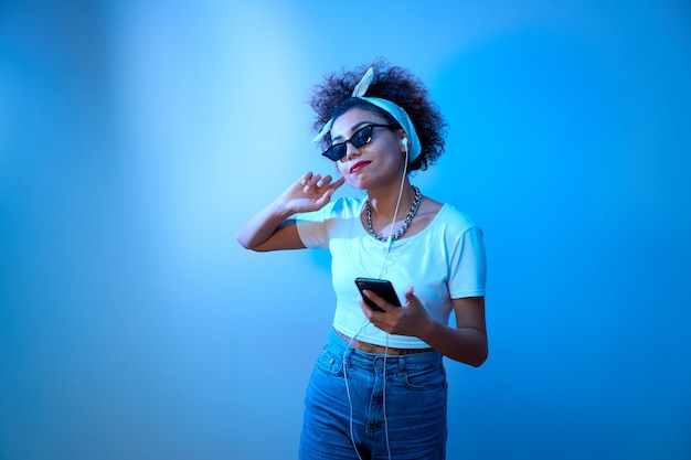 Une fille à la mode avec des boucles afro écoute et apprécie la musique avec des écouteurs à la lumière du néon bleu, des danses de modèle de style hip hop et se détendre