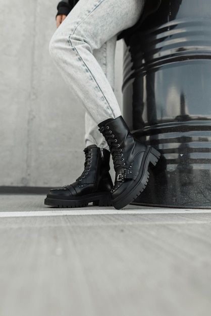 Fille à la mode dans des chaussures et des jeans en cuir noir à la mode se tient près d'un tonneau en métal noir dans la rue