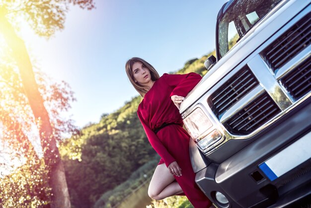 Fille de mode en robe rouge élégante, debout près de sa voiture moderne et se détendre dans la nature