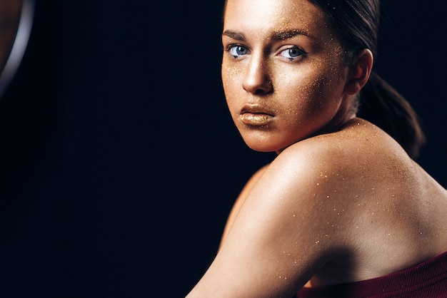 Fille de modèle d'art de mode de beauté avec la peau d'or. Bouchent portrait de fille aux yeux bleus sur fond noir