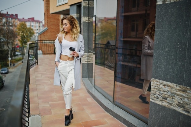 Fille modèle blonde bouclée élégante porter sur blanc avec une tasse de café à portée de main posant contre une grande fenêtre