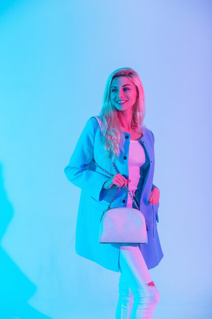 Fille modèle blonde heureuse avec un sourire blanc portant des vêtements élégants à la mode avec un blazer bleu, un débardeur blanc et un jean tenant un sac blanc dans le studio sur un néon rose de couleur pastel