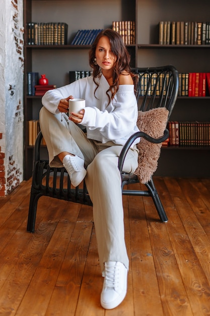 Une fille modèle dans une veste blanche est assise sur une chaise avec une tasse de café dans ses mains