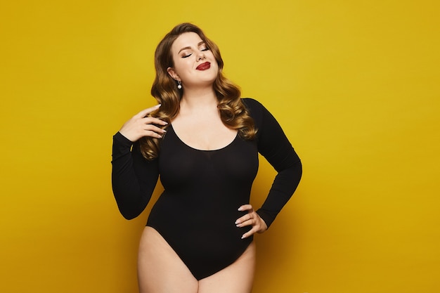 Fille de modèle grande taille avec un maquillage lumineux en body noir posant sur le fond jaune. Jeune femme de taille plus en body noir isolé sur fond jaune