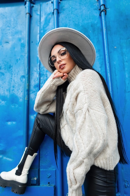 Fille de modèle de mode avec des lunettes vintage en chandail tricoté à la mode pantalon en cuir chapeau et bottes cool pose près d'un fond en métal bleu coloré