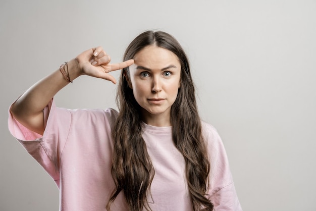 La fille montre son doigt sur sa tempe Fille brune aux cheveux longs sur fond gris