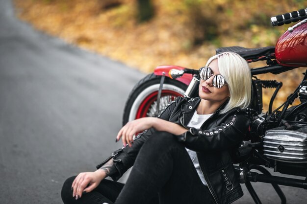 Fille de motard jolie blonde à lunettes de soleil assis près de moto rouge sur la route dans la forêt