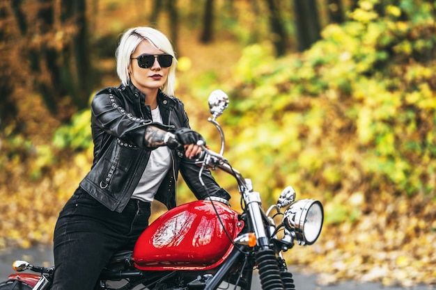 Fille de motard jolie blonde à lunettes de soleil avec moto rouge sur la route dans la forêt