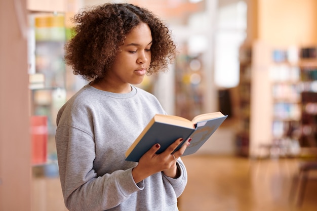 Fille multiculturelle aux cheveux ondulés en sweat-shirt gris lecture livre de contes dans la bibliothèque de l'université