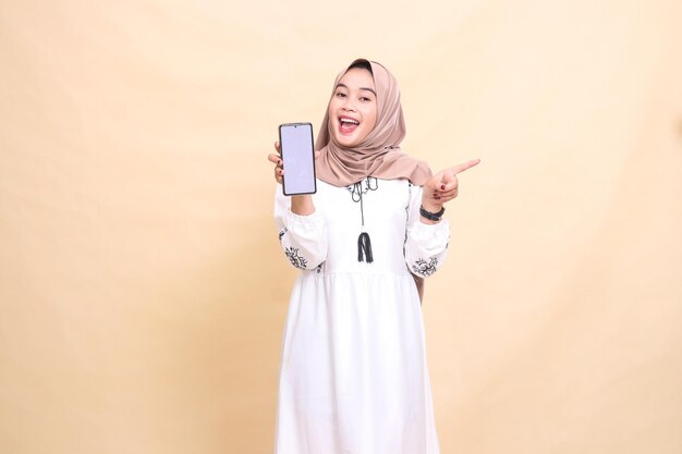 Une fille musulmane asiatique adulte portant un hijab sourit heureusement en montrant un écran de téléphone portable pointing