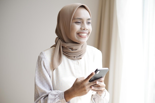 Une fille musulmane asiatique en vêtements décontractés utilise un smartphone et sourit tout en se tenant près de la fenêtre
