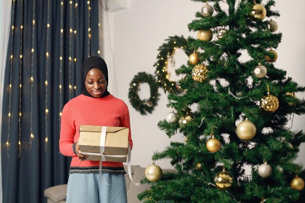 Fille musulmane en hijab noir debout près de l'arbre de Noël avec boîte-cadeau